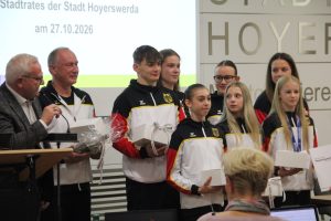 Oberbürgermeister Torsten Ruban-Zeh spricht am Rednerpult. Neben ihm stehen Trainer Sergej Jeriomkin und sieben Sportakrobatinnen und Sportakrobaten in schwarz-rot-weißen Trainingsanzügen. Mehrere halten Geschenkverpackungen, zwei tragen Medaillen.