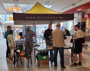 Mehrere Menschen stehen in kleinen Gesprächsgruppen an einem Messestand mit einem gelben Dach und der Aufschrift „www.darumwhy.de“. Auf einem Tisch liegen Informationsmaterialien.