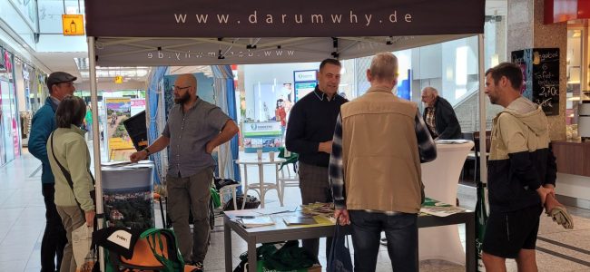 Mehrere Menschen stehen in kleinen Gesprächsgruppen an einem Messestand mit einem gelben Dach und der Aufschrift „www.darumwhy.de“. Auf einem Tisch liegen Informationsmaterialien.