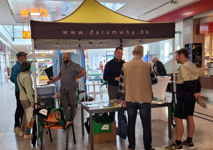 Mehrere Menschen stehen in kleinen Gesprächsgruppen an einem Messestand mit einem gelben Dach und der Aufschrift „www.darumwhy.de“. Auf einem Tisch liegen Informationsmaterialien.