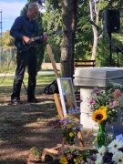 Ein Mann spielt im Freien E-Gitarre. Neben ihm stehen der Gedenkstein, ein gerahmtes Porträt und Blumen.