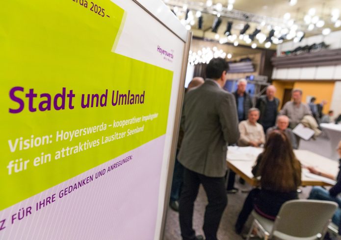 Bildbeschreibung: Im Vordergrund ein Plakat mit der Überschrift „Stadt und Umland – Vision: Hoyerswerda – kooperativer Impulsgeber für ein attraktives Lausitzer Seenland". Im Hintergrund stehen und sitzen mehrere Personen an Stelltischen mit ausgebreiteten Plänen in einem Saal mit Bühnenbeleuchtung. Alternativtext: Im Vordergrund ein Plakat mit der Überschrift „Stadt und Umland – Vision: Hoyerswerda – kooperativer Impulsgeber für ein attraktives Lausitzer Seenland". Im Hintergrund stehen und sitzen mehrere Personen an Stelltischen mit ausgebreiteten Plänen in einem Saal mit Bühnenbeleuchtung. Beschriftung mit Bildnachweis: Bürgerwerkstatt Hoyerswerda. Foto: G. Menzel