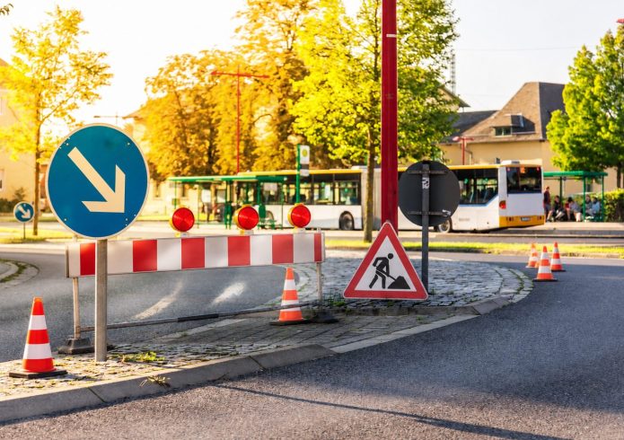 Eine rot-weiße Straßensperre mit drei Warnleuchten blockiert eine Fahrspur. Daneben stehen ein Baustellenschild, mehrere orange-weiße Leitkegel und ein blaues Richtungsschild. Im Hintergrund halten ein Stadtbus und wartende Personen an einer Haltestelle. Das Bild wurde mit KI bearbeitet.