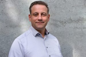 Porträt von Christian Hoffmann in einem hellen, gemusterten Hemd vor einer grauen Betonwand.