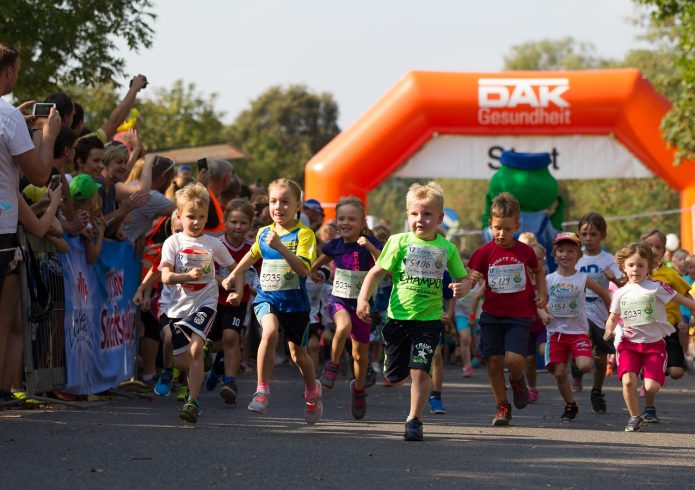 Eine Gruppe von Kindern mit Startnummern läuft über eine Straße durch ein aufblasbares Starttor mit der Aufschrift „DAK Gesundheit, Start". Am Streckenrand feuern zahlreiche Zuschauende die Kinder an.