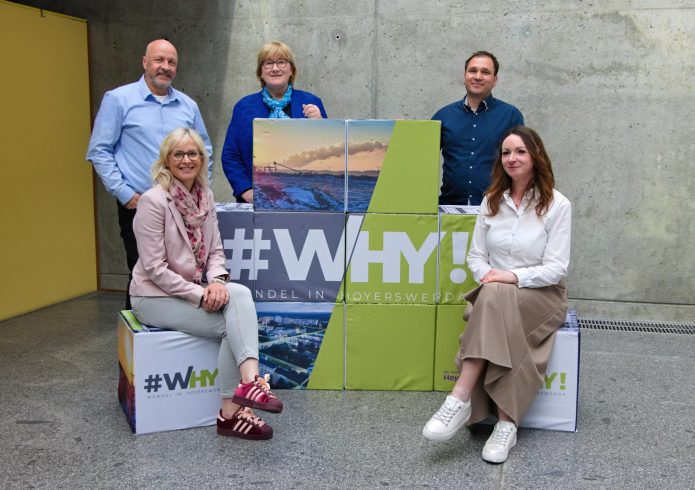 Fünf Mitglieder des Projektteams Strukturwandel vor einer Betonwand. Zwei sitzen, drei stehen um gestapelte Würfel mit Stadtmotiven und der Aufschrift „#WHY! – Wandel in Hoyerswerda“.