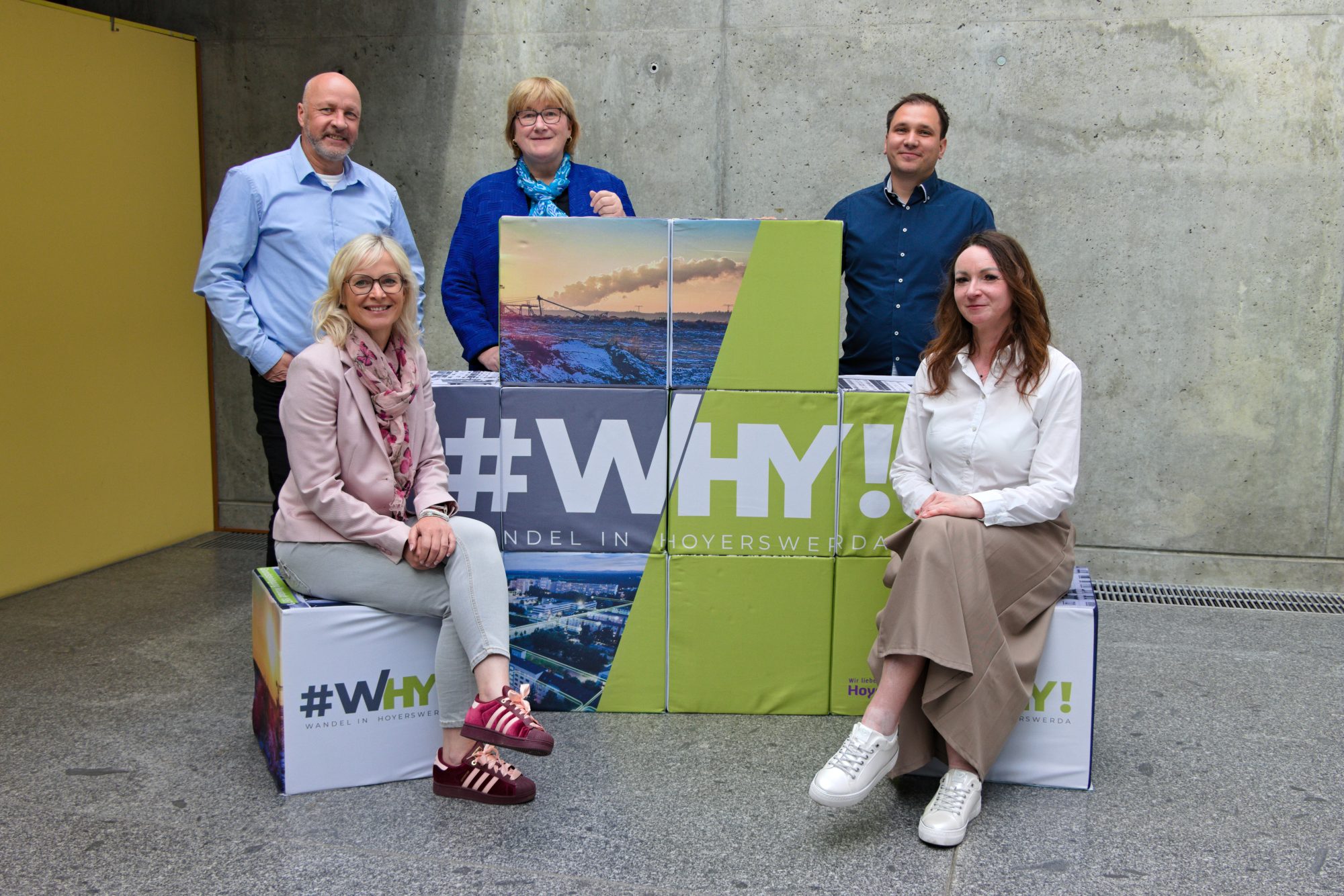 Fünf Mitglieder des Projektteams Strukturwandel vor einer Betonwand. Zwei sitzen, drei stehen um gestapelte Würfel mit Stadtmotiven und der Aufschrift „#WHY! – Wandel in Hoyerswerda“.