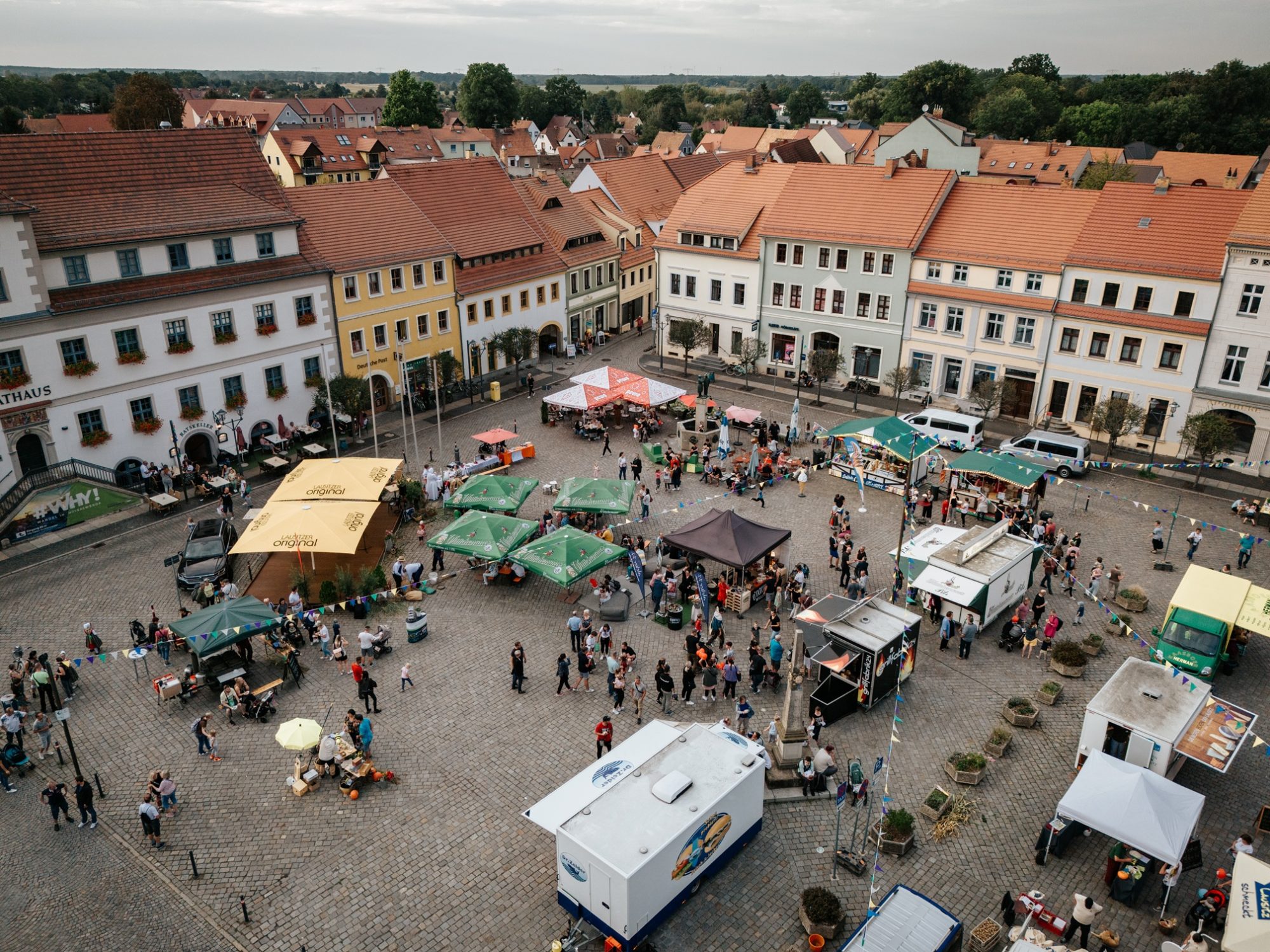 Luftaufnahme des Marktplatzes von Hoyerswerda mit Rathaus, umliegenden Altstadthäusern und einem Brunnen. Auf dem Platz stehen zahlreiche Verkaufsstände, Imbisswagen, Sonnenschirme und Pavillons, umgeben von vielen Menschen.