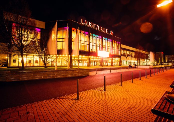 Nächtliche Außenansicht der Lausitzhalle mit beleuchteter Glasfassade und Schriftzug „Lausitzhalle" auf dem Dach. Davor eine breite Promenade mit Bänken und Straßenlaternen.