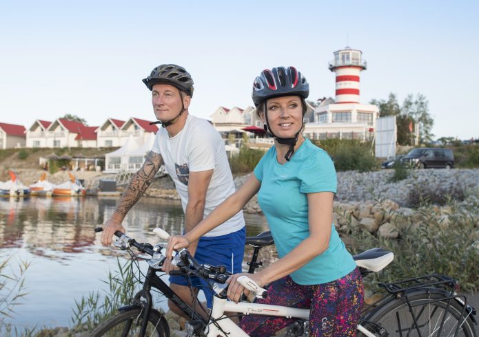 Ein Mann und eine Frau mit Fahrradhelmen schieben ihre Fahrräder am Ufer des Geierswalder Sees, im Hintergrund das rot-weiß gestreifte Leuchtturm-Hotel und Ferienhäuser.
