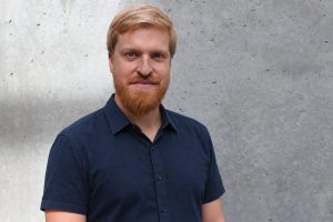 Porträt von Sebastian Schindler mit rötlichem Bart und dunkelblauem Hemd vor einer grauen Betonwand.