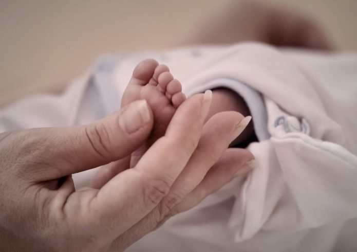 Nahaufnahme: Eine erwachsene Hand hält sanft den Fuß eines Neugeborenen, das in eine helle Decke gewickelt ist.
