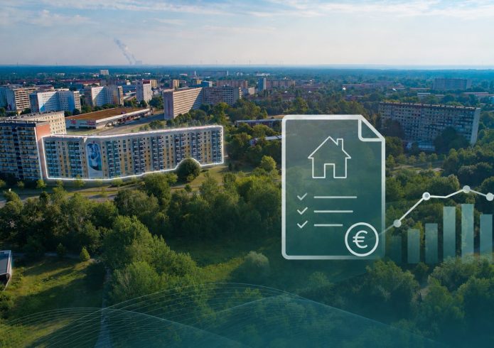 Luftaufnahme eines Wohngebiets in Hoyerswerda mit mehreren Wohnblocks und Grünflächen. Im Vordergrund überlagert eine grafische Darstellung eines Dokuments mit Haussymbol, Checklisten, Eurozeichen und einem ansteigenden Balkendiagramm.