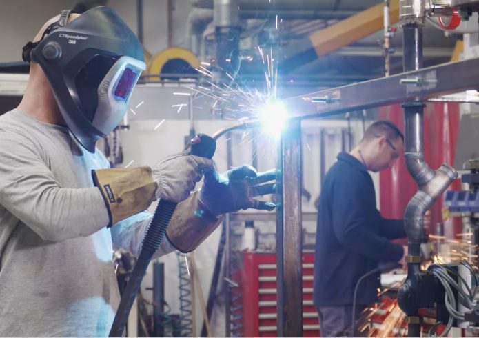 Ein Schweißer in Schutzausrüstung und mit Schweißmaske bearbeitet ein Metallgestell, Funken sprühen. Im Hintergrund arbeitet ein weiterer Mitarbeiter an einer Rohrkonstruktion in einer Werkshalle.