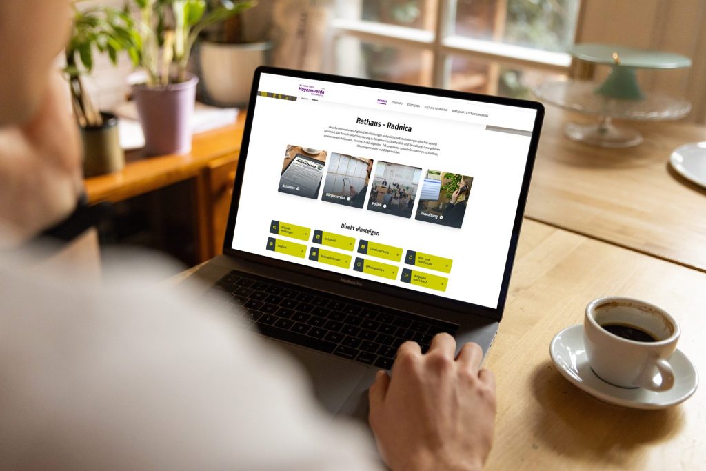 Bildmontage: Eine Person sitzt an einem Holztisch vor einem Laptop. Auf dem Bildschirm ist der Bereich „Rathaus“ der Website hoyerswerda.de zu sehen. Neben dem Laptop steht eine Kaffeetasse.