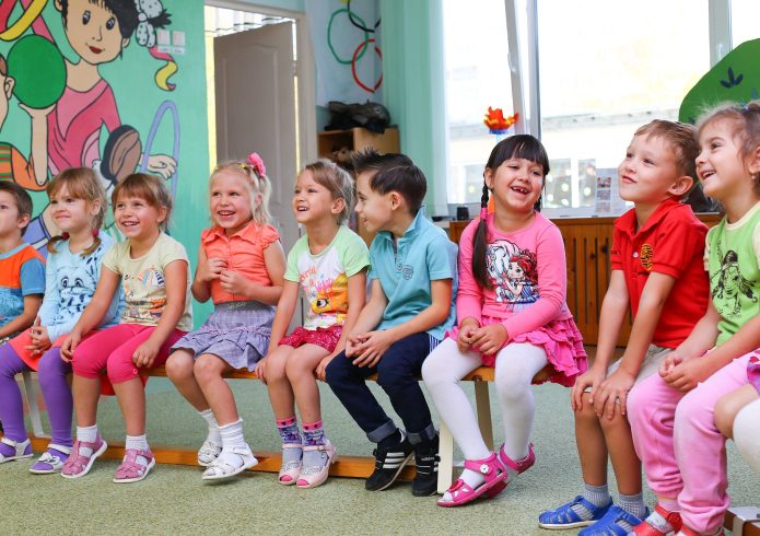 Eine Gruppe von Kindern sitzt lachend auf einer Bank in einem bunt gestalteten Gruppenraum einer Kindertagesstätte.