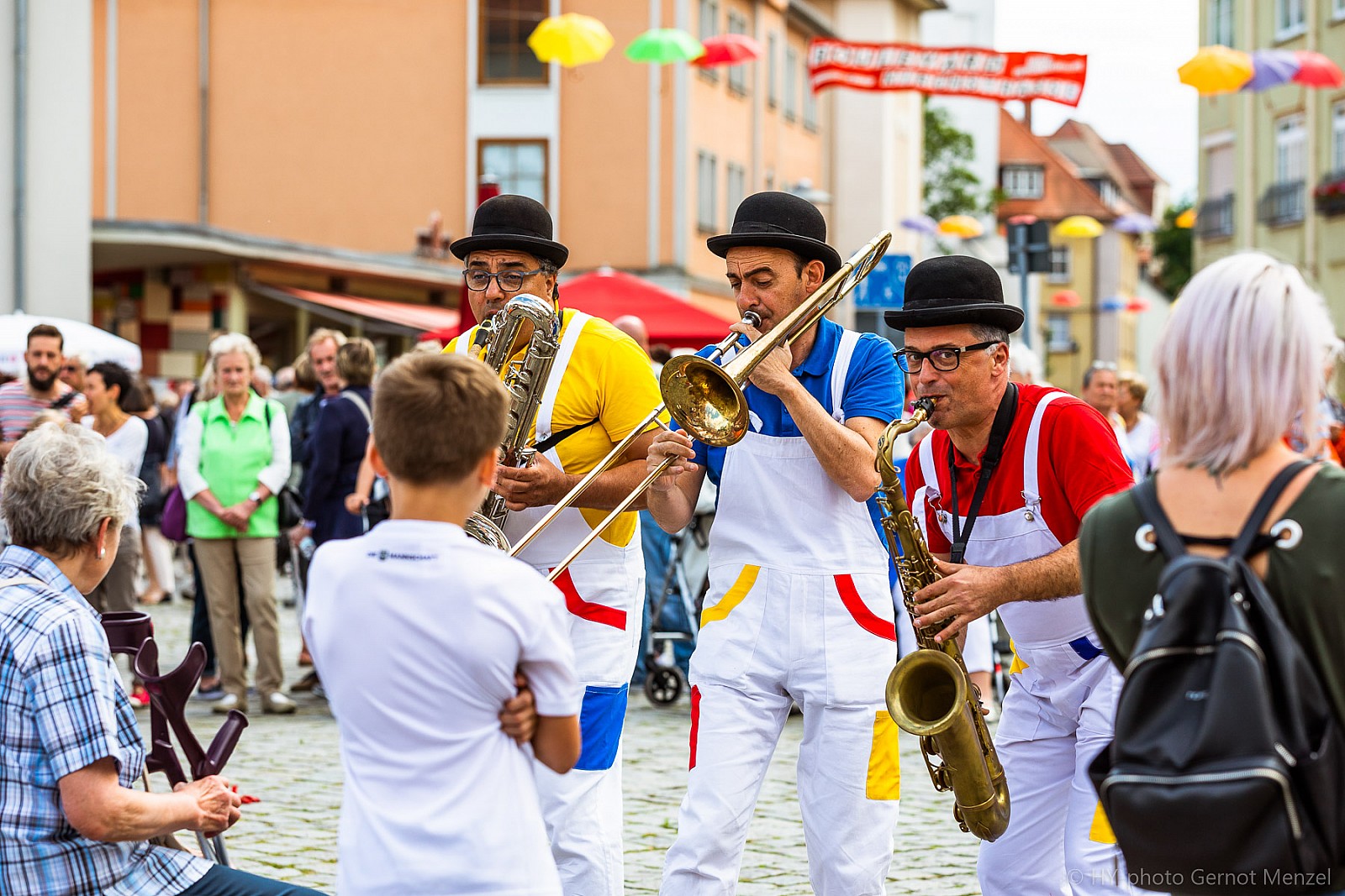 Drei Musiker in Melonenhüten und bunten Latzhosen spielen mit Saxophon und Posaune vor Publikum auf einem Straßenfest, im Hintergrund bunte Sonnenschirme und Häuserfassaden.